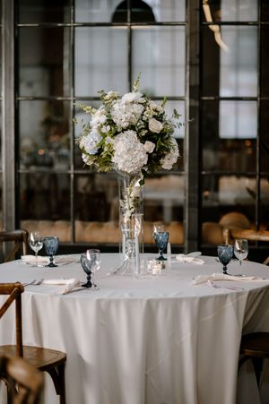 Elegant wedding reception tables with white tablecloths and floral centerpieces in tall glass vases at an indoor venue.