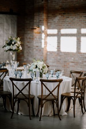 Elegant wedding reception table with white flowers, rustic wooden chairs and exposed brick walls in an industrial venue.