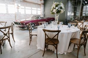 Elegant wedding reception table with white linens and floral centerpiece next to classic burgundy convertible car in industrial venue.