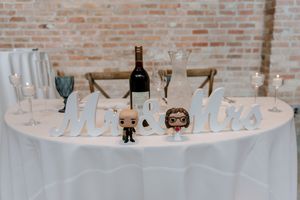 White wedding table with Mr & Mrs sign, wine bottle, glasses, and pop culture figurines against brick wall backdrop.