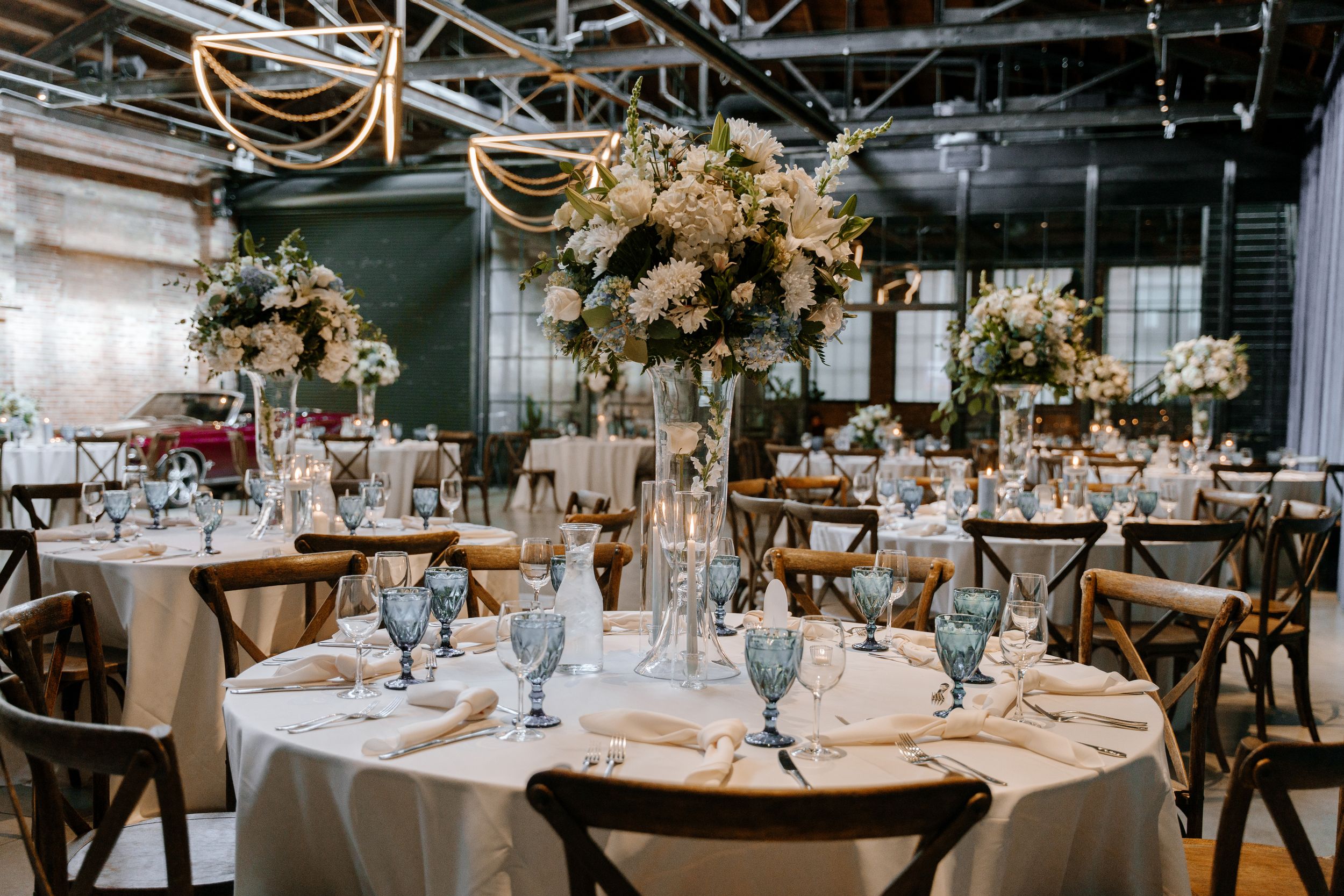 Elegant wedding reception tables with blush floral centerpieces and cross-back chairs in an industrial venue setting.