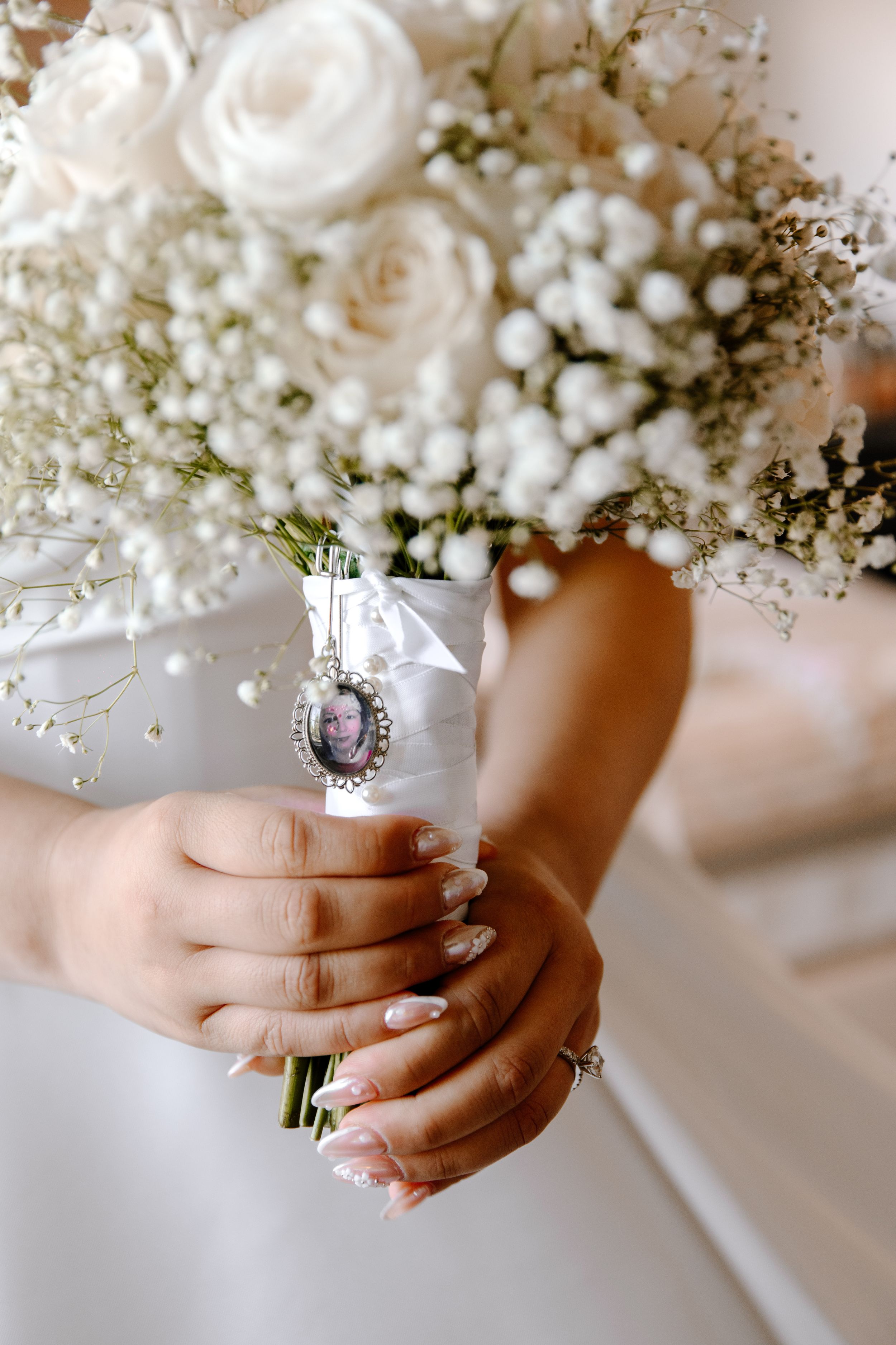 Manicured hands hold an elegant white bridal bouquet with delicate baby's breath and roses against a light background.