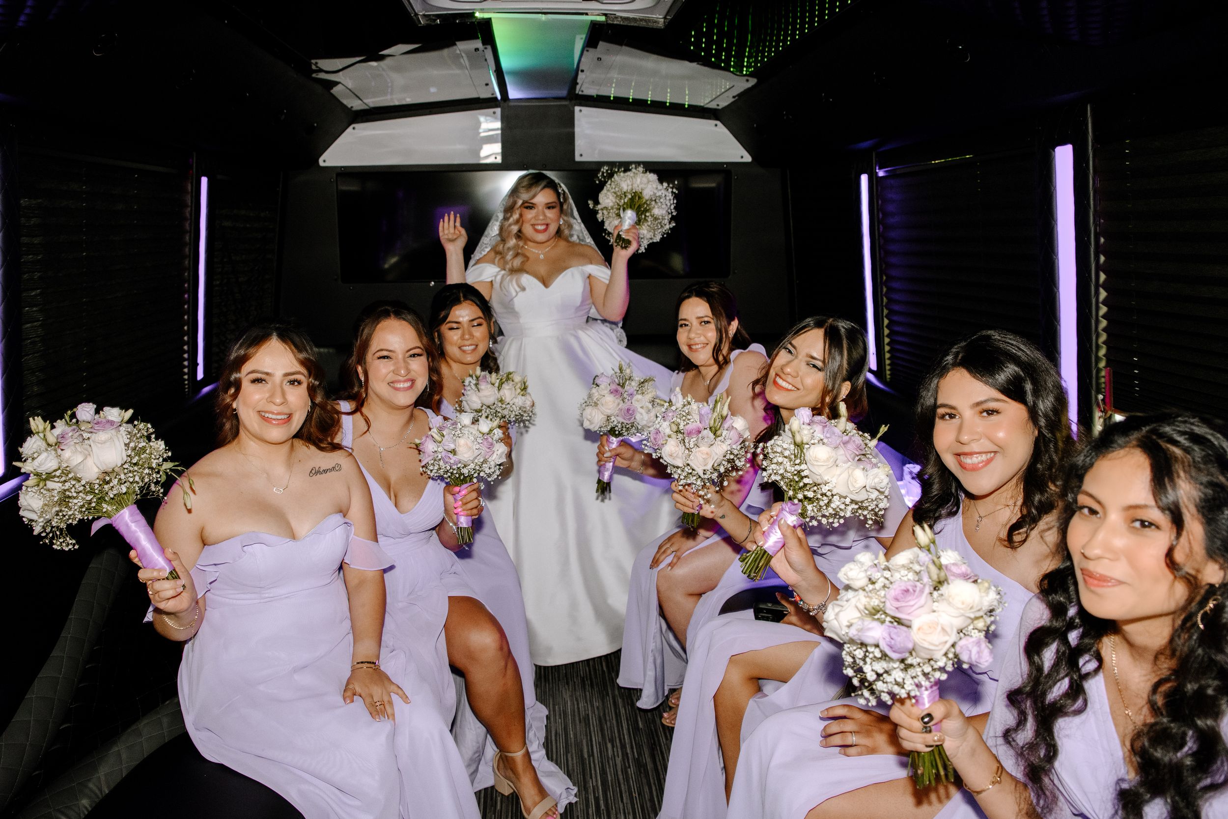 Bridal party in light purple bridesmaid dresses holding floral bouquets pose together inside a party bus.
