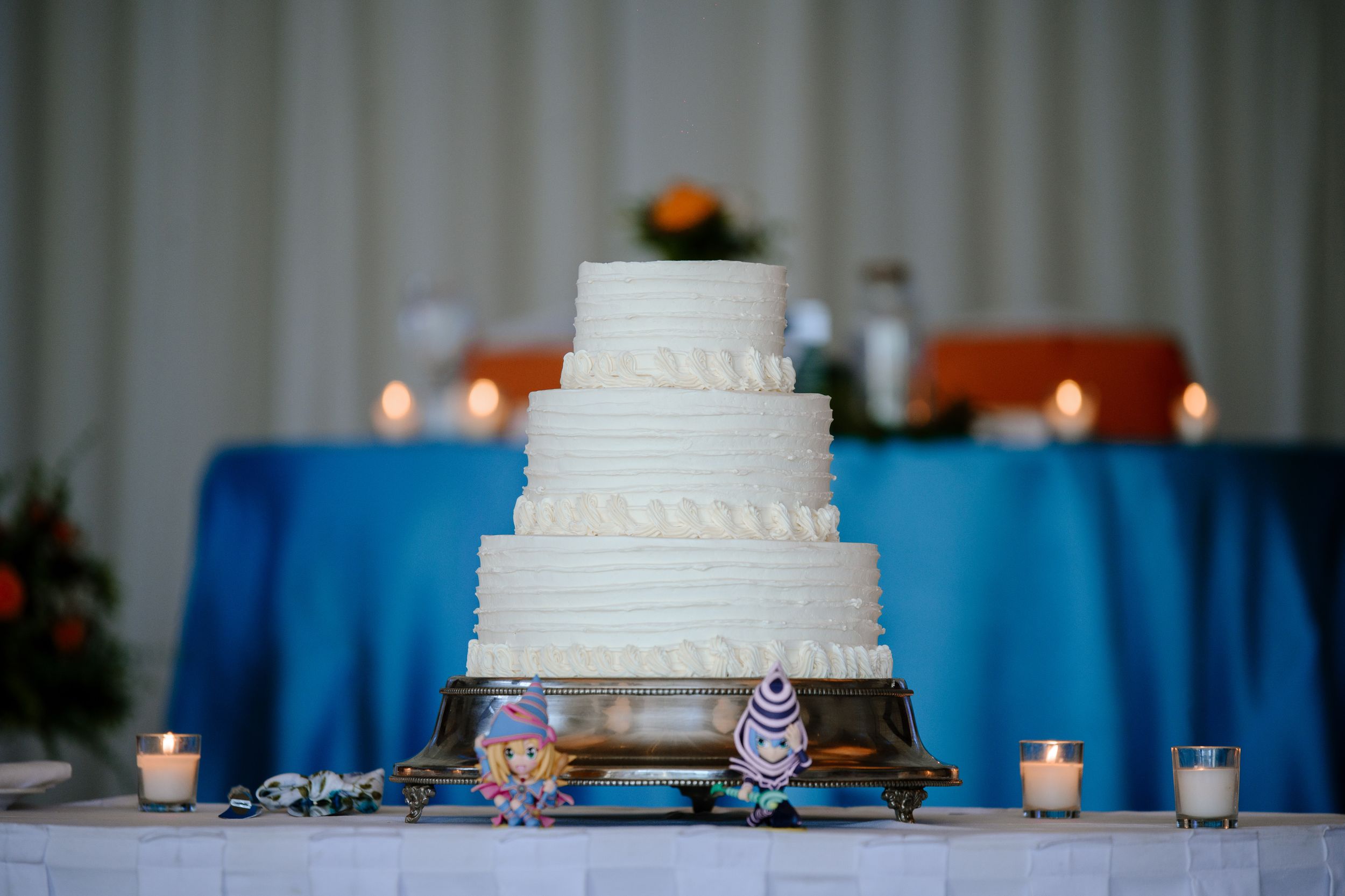 A white tiered wedding cake with ornate designs sits on a table with blue linens and glowing candles.