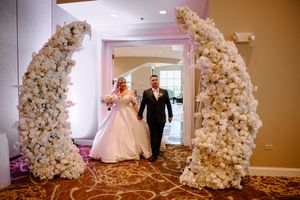 A wedding dress glides through a hallway decorated with white floral arrangements and delicate lighting.