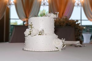 A two-tiered white wedding cake decorated with delicate silver accents sits on a table with draped curtains in the background.