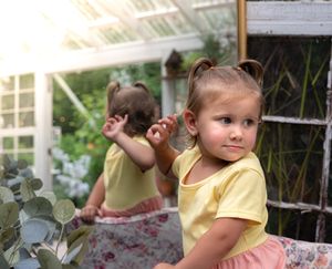 A child in a yellow and coral dress poses thoughtfully in a garden setting.