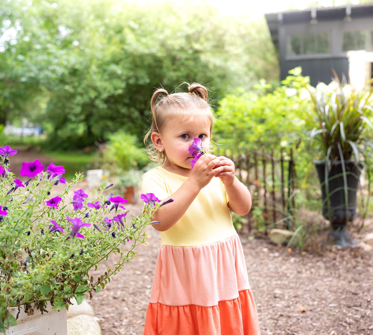 A child in a tiered dress smells purple flowers in a beautiful garden setting.