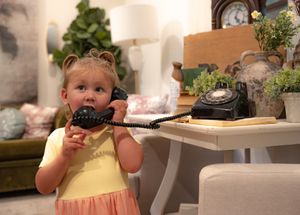 A child in a yellow dress playfully holds a vintage rotary telephone in an indoor setting.