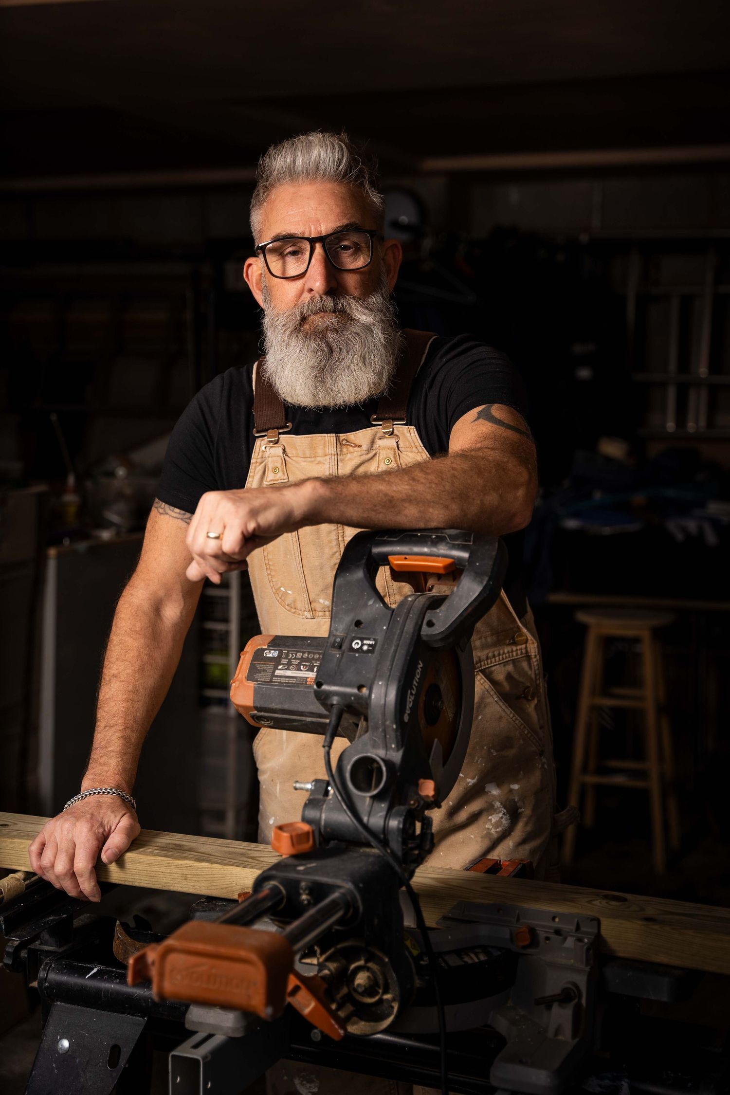A Winchester based small business owner poses in his workshop during a personal branding photoshoot. He wears overalls and leans on a saw.