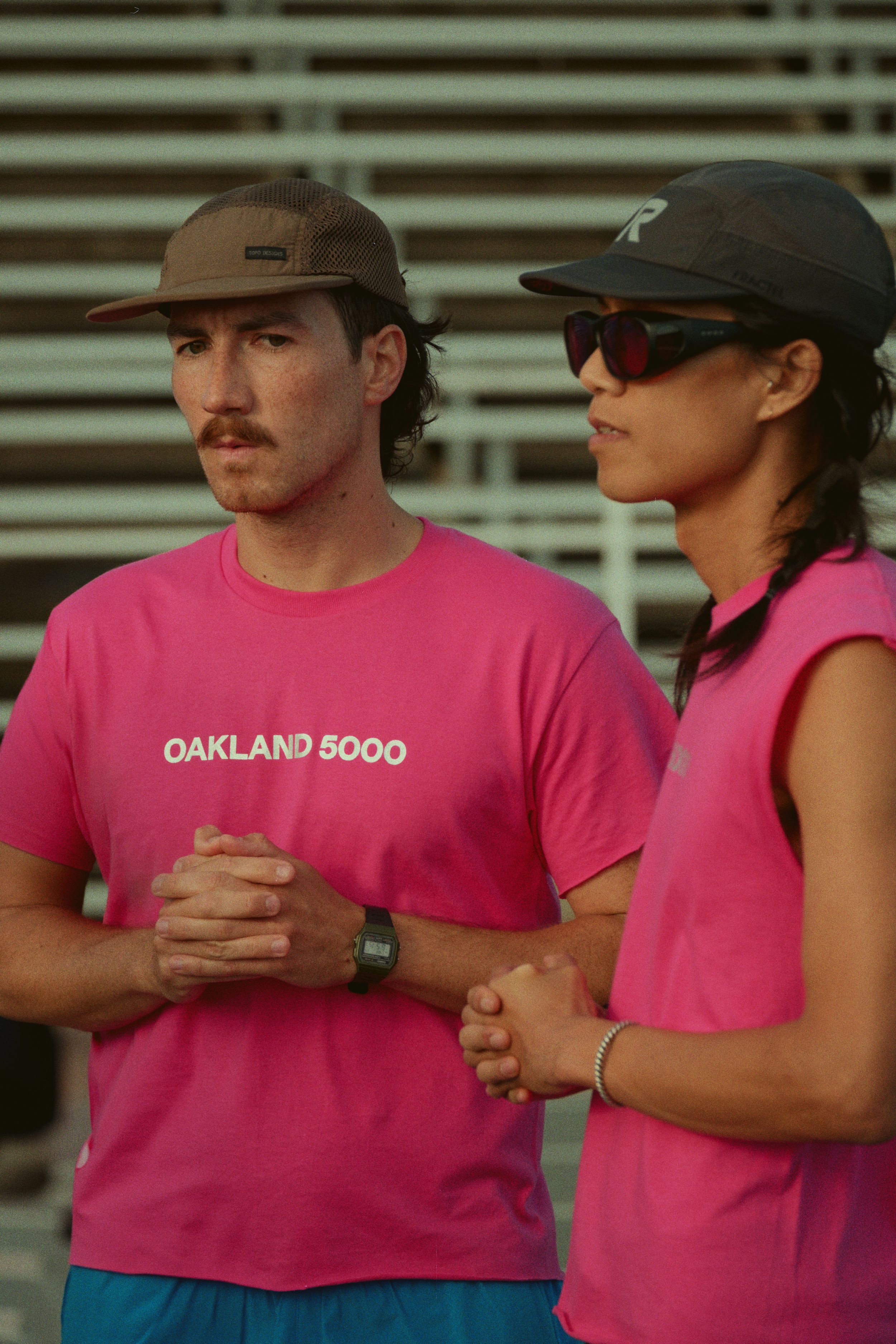 Two people wearing pink t-shirts and baseball caps stand together in front of stadium bleachers.