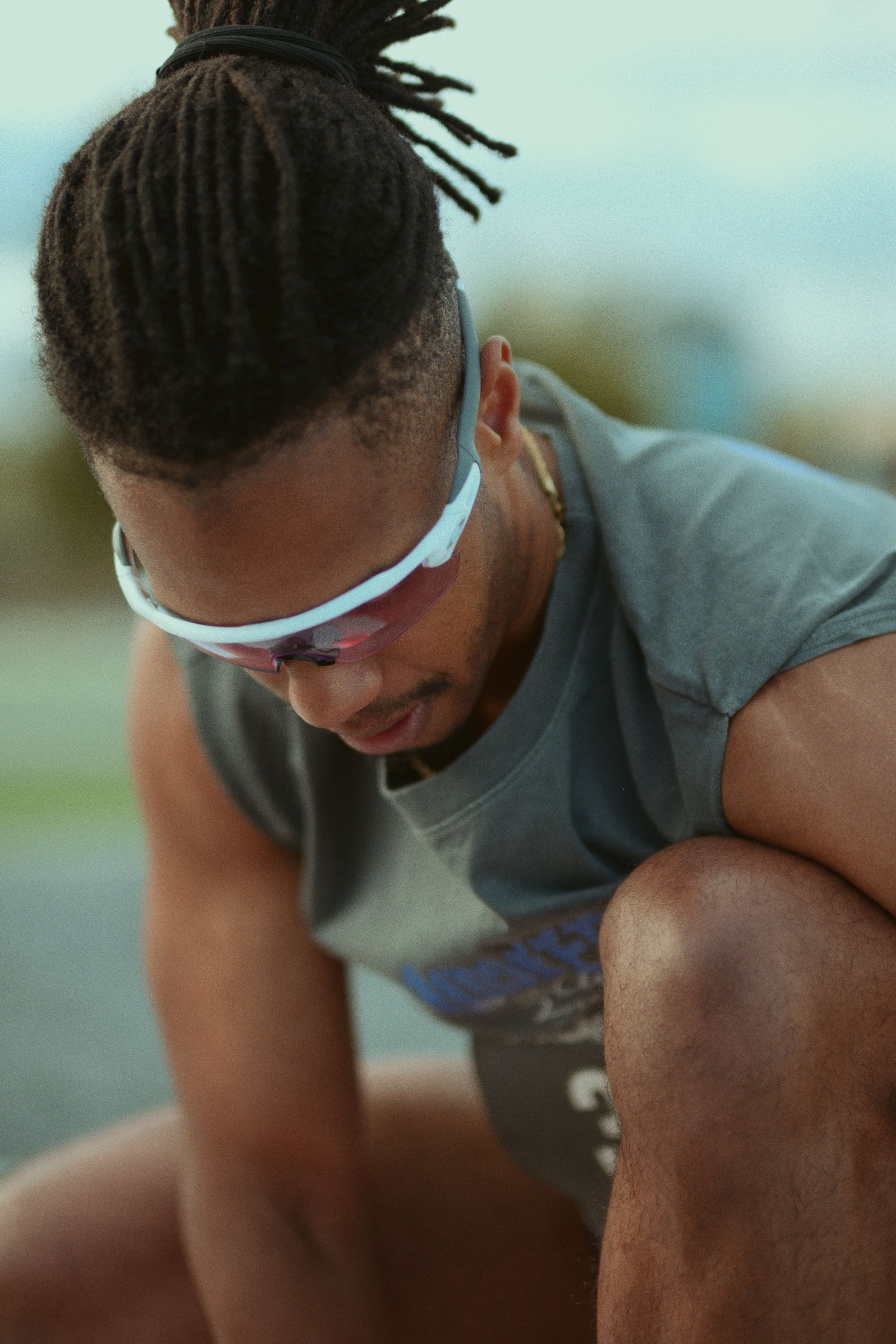 Athletic person in a gray t-shirt wearing white sunglasses and dreadlocks in a top knot hairstyle rests outdoors.