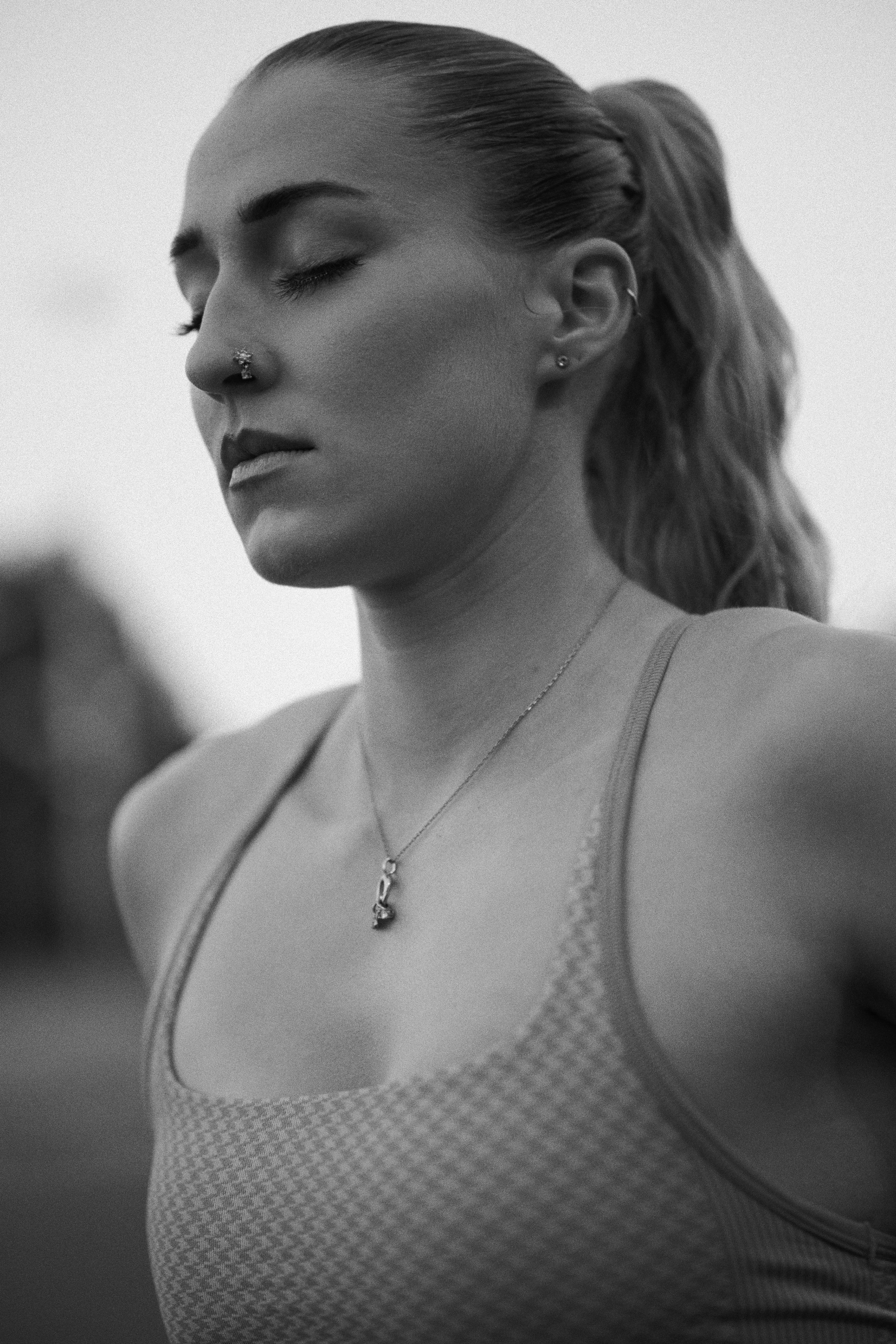 Black and white portrait of someone with a ponytail and nose piercing, wearing a textured tank top and necklace.