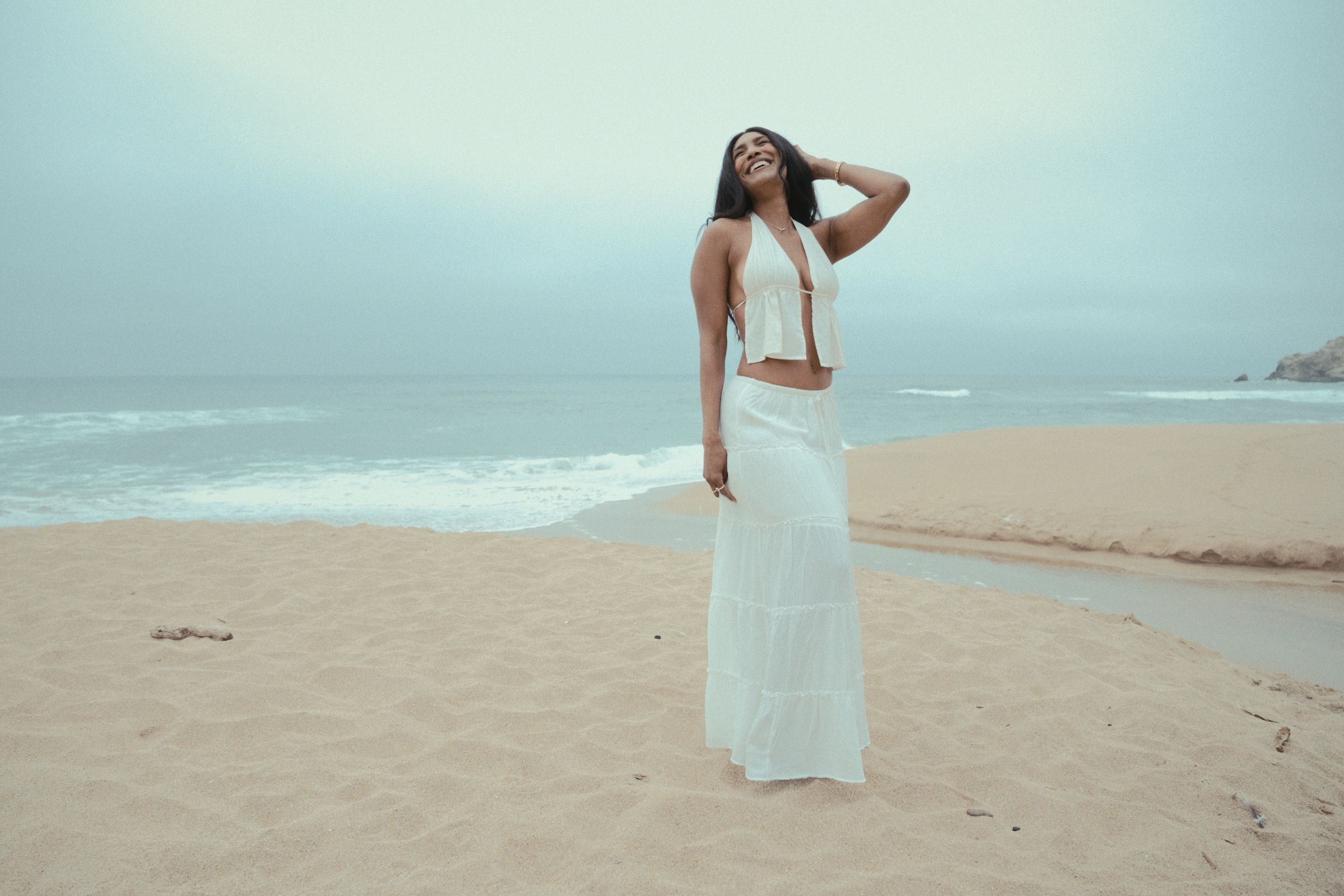 Someone wearing a flowing white two-piece outfit poses elegantly on a misty beach by the ocean.