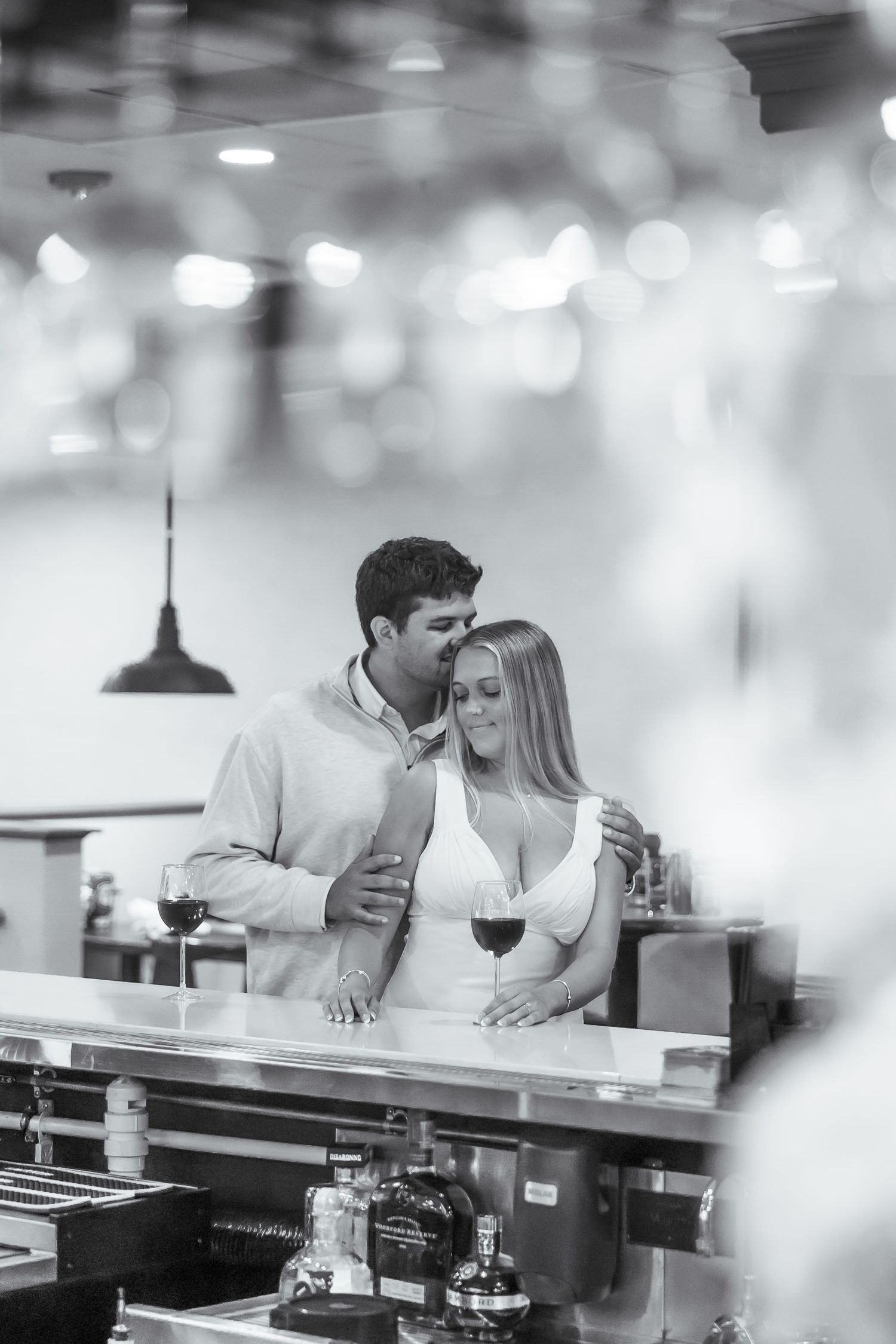 Black and white romantic photo of couple sharing intimate moment at restaurant table with wine glasses and bokeh lights.
