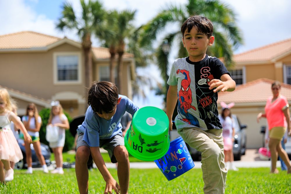 Children playing outdoor egg hunt game on green grass, boy in Spider-Man shirt carrying colorful buckets on sunny day.