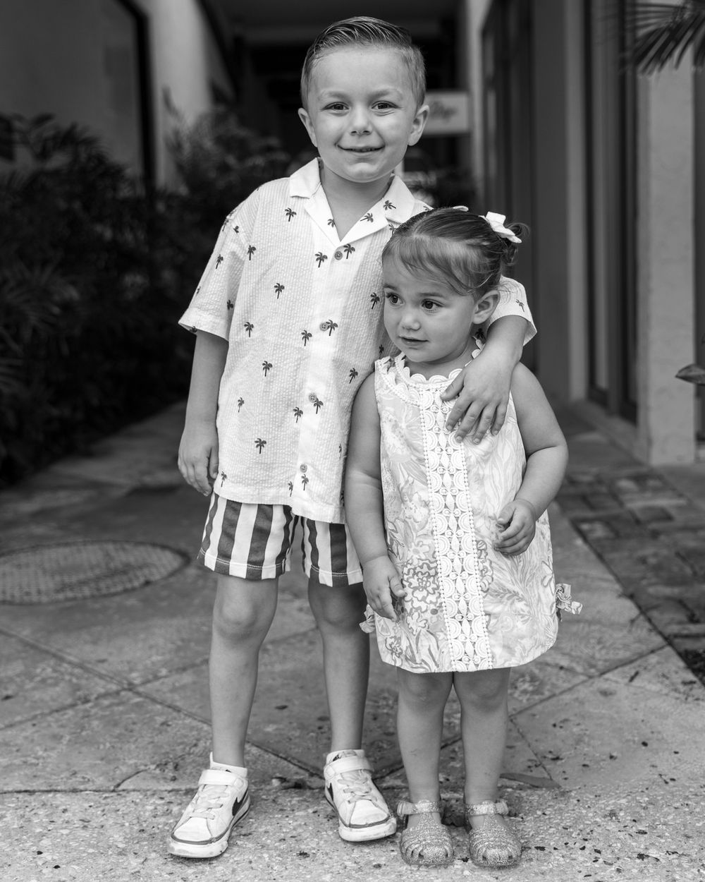 Two young siblings pose together outdoors, boy in striped shorts hugging little girl in white dress, black and white photo.
