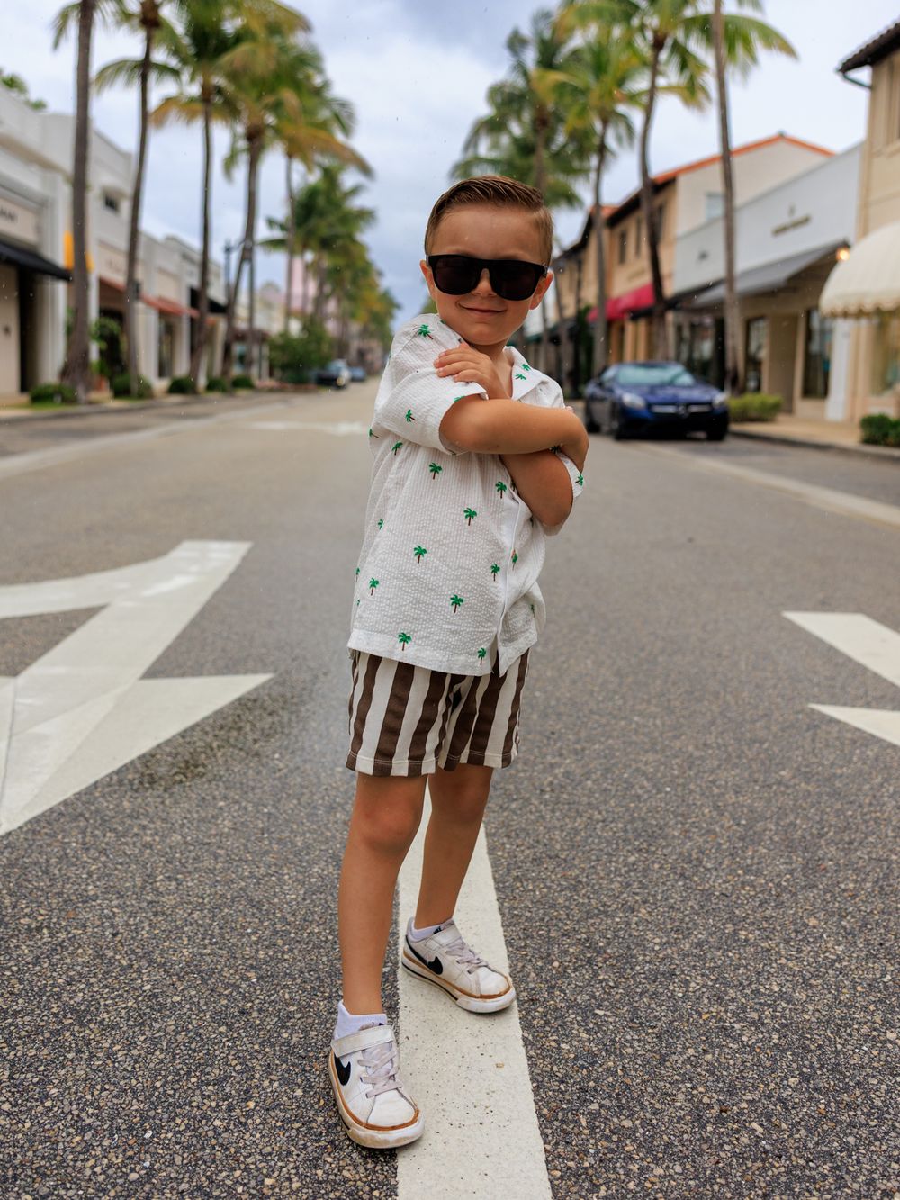 A stylish young boy in sunglasses, palm tree shirt, striped shorts, and sneakers poses confidently on a sunny street.