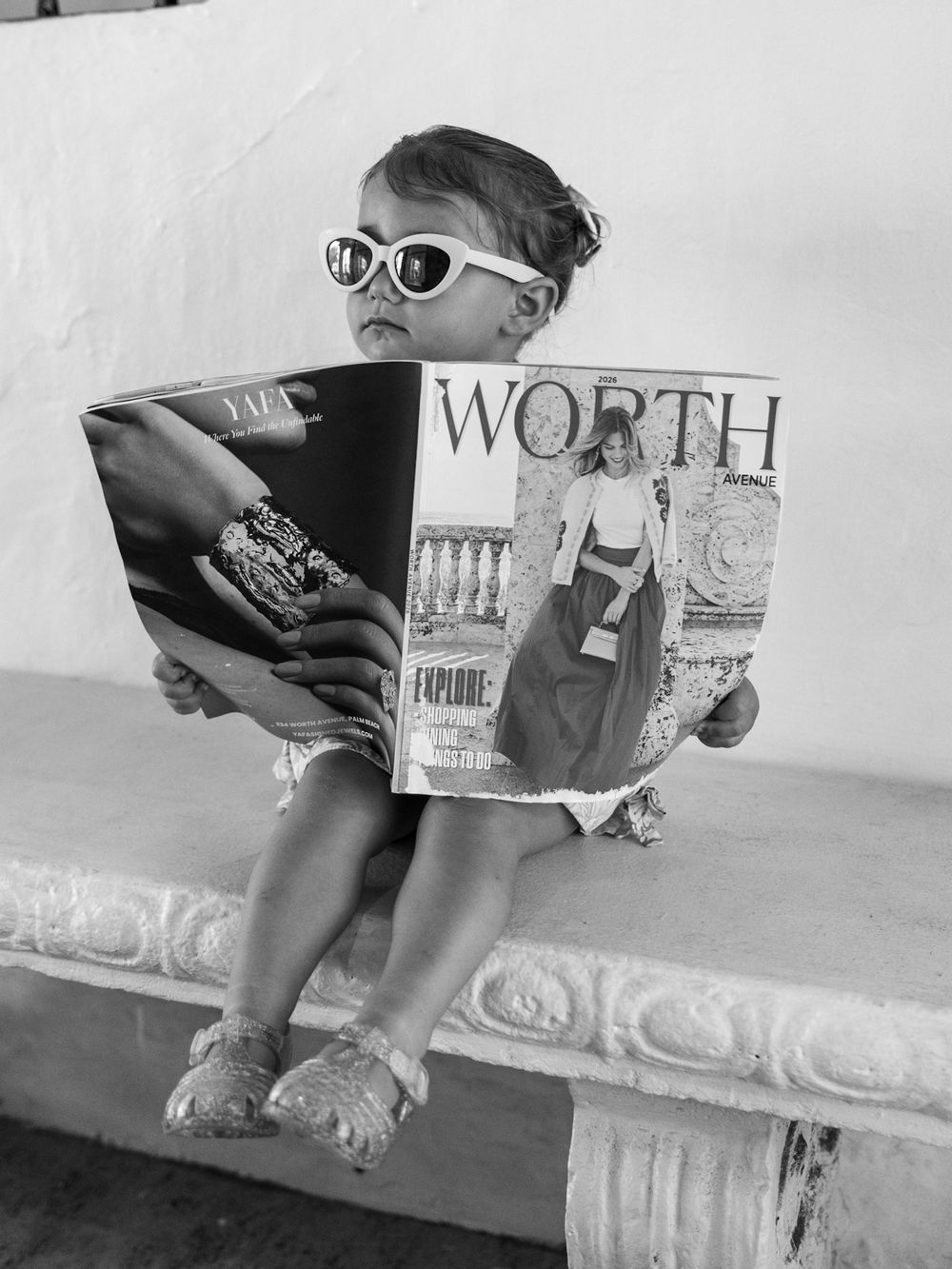 A toddler wearing sunglasses sits on a bench reading Worth magazine in a charming black and white photo.