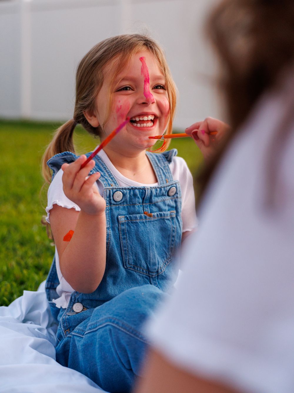 Child in denim overalls laughs joyfully while having face paint applied during outdoor activity on grass.