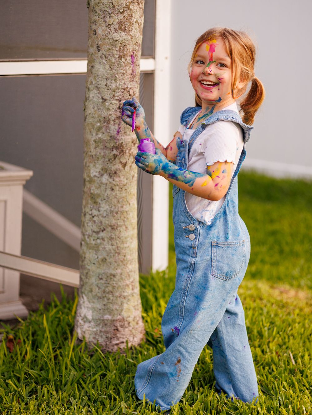 A child in denim overalls plays in the grass near a white porch post, showing a series of joyful movements.