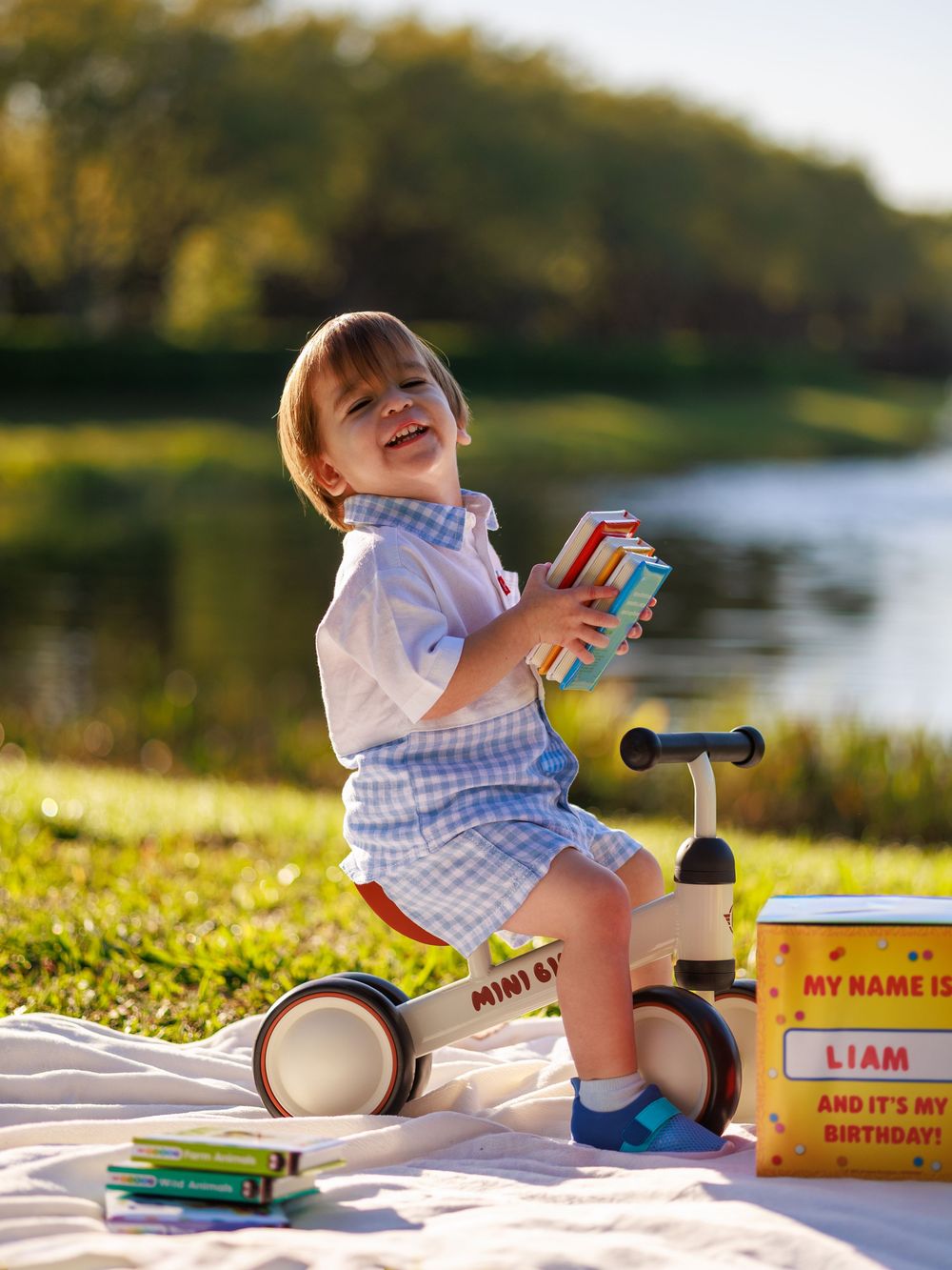 A toddler in blue and white outfit sits on a small tricycle outdoors by a lake during golden hour.