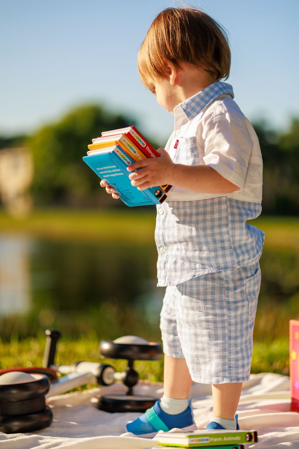 Small child in gingham overalls holds books while standing outdoors in warm sunlight.