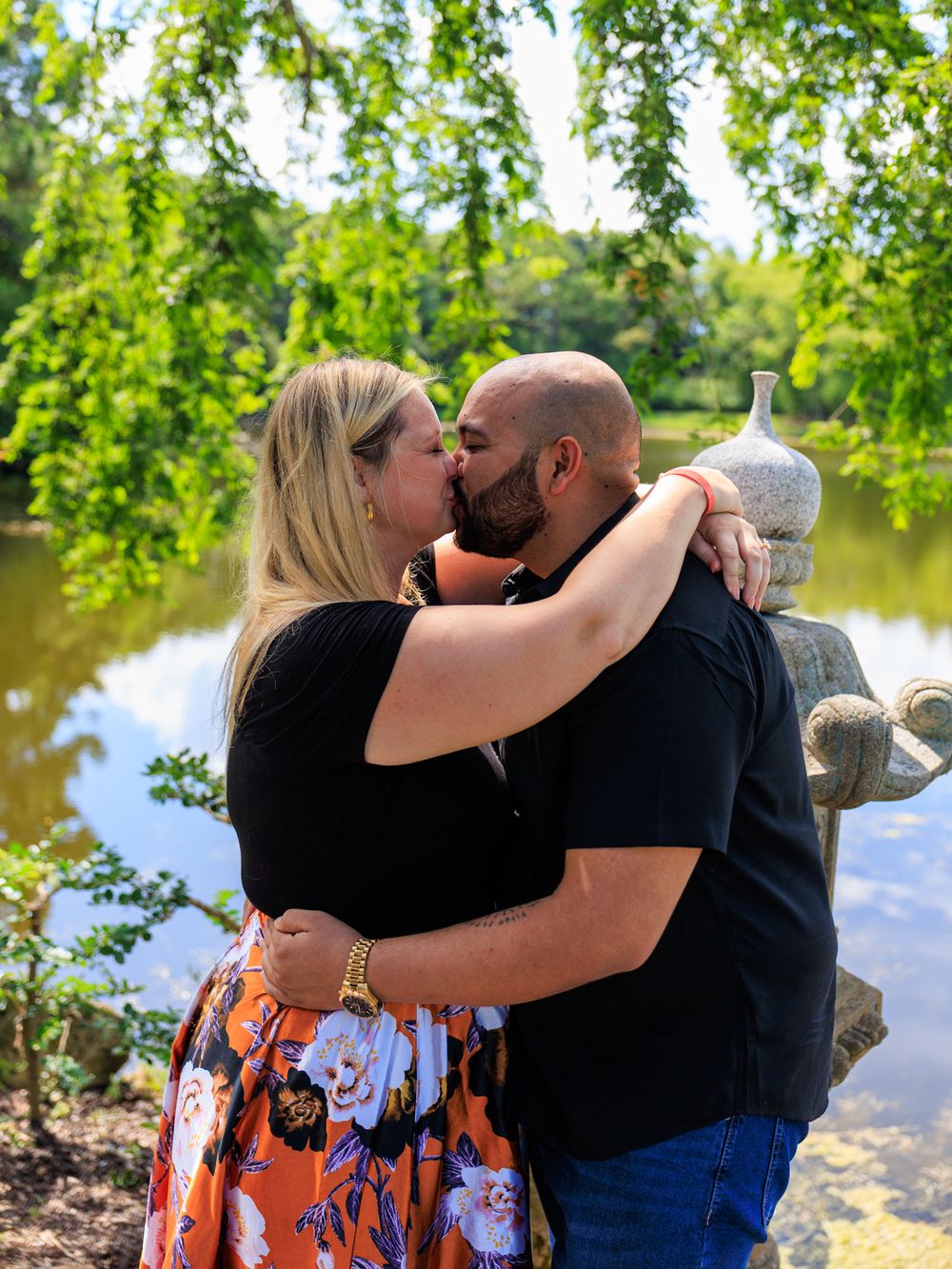 Couple sharing a romantic kiss by a serene pond surrounded by lush green trees in a garden setting.