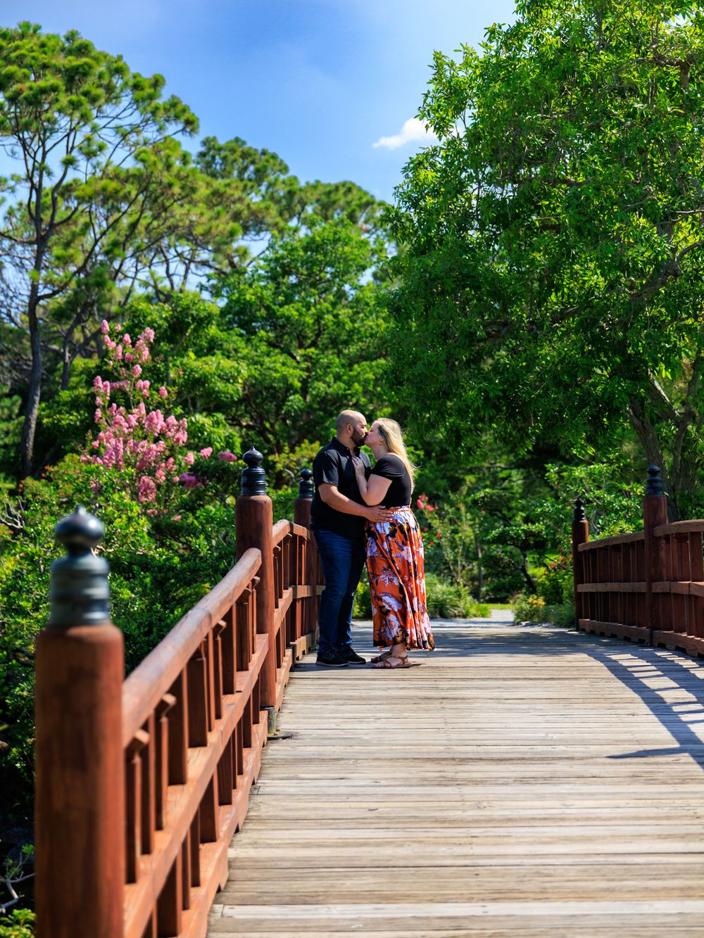 Couple kissing on a wooden bridge surrounded by vibrant greenery and blooming flowers on a sunny day.