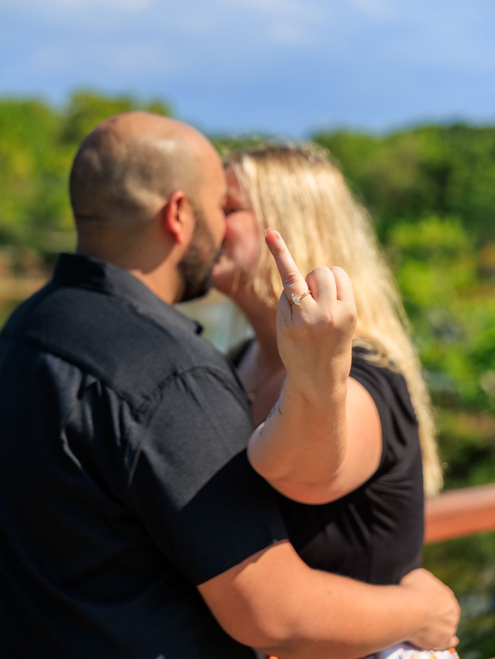 Couple kissing outdoors while woman shows off engagement ring, with lush green trees and blue sky background.