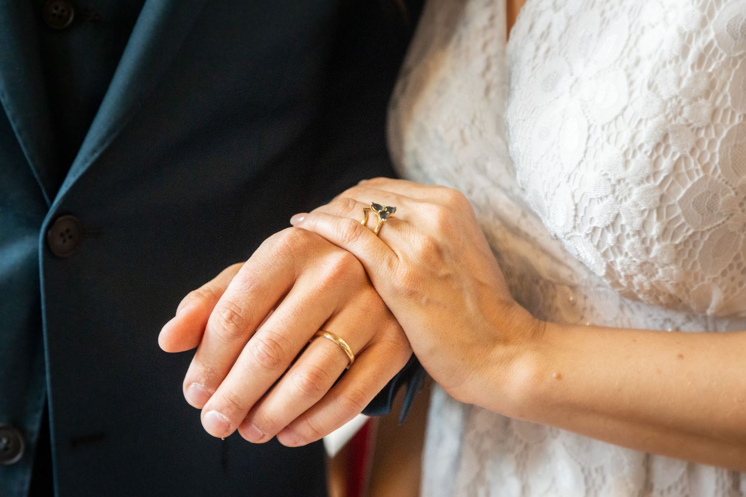 Close-up photo of newlyweds' hands showing wedding rings, with white dress and black suit in background.