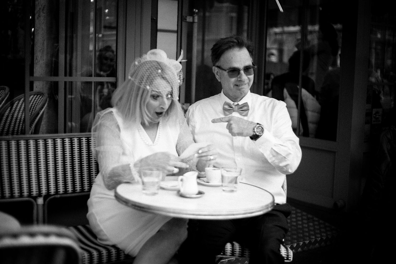 A stylish couple laughs together while enjoying coffee at an outdoor Parisian cafe table in black and white.