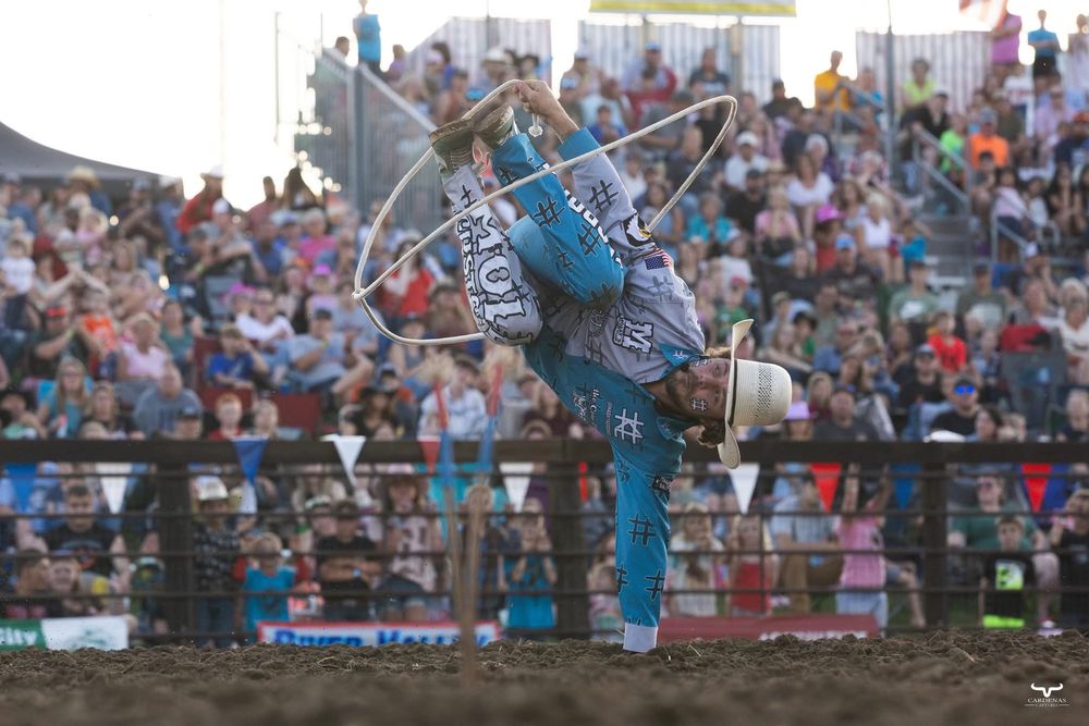 Crowd watches rodeo athlete perform dangerous maneuvers on bucking bronco at packed stadium event.