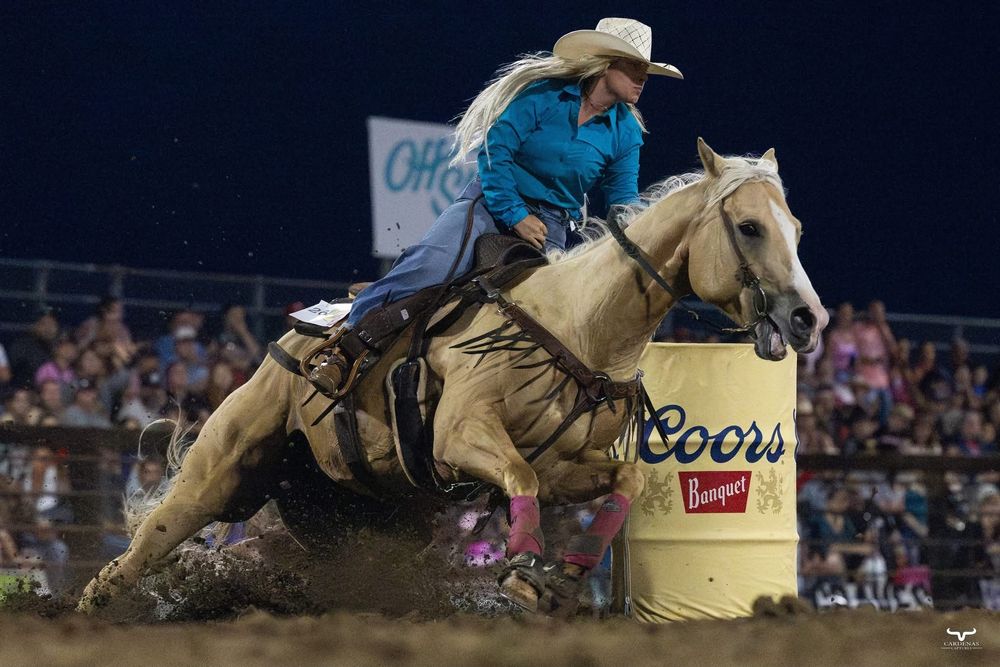 A barrel racer on a white horse makes a sharp turn around a barrel during a nighttime rodeo competition.