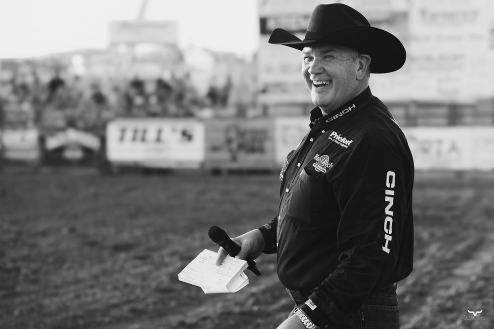 A rodeo official wearing a black cowboy hat and jacket stands in a dirt arena holding papers.