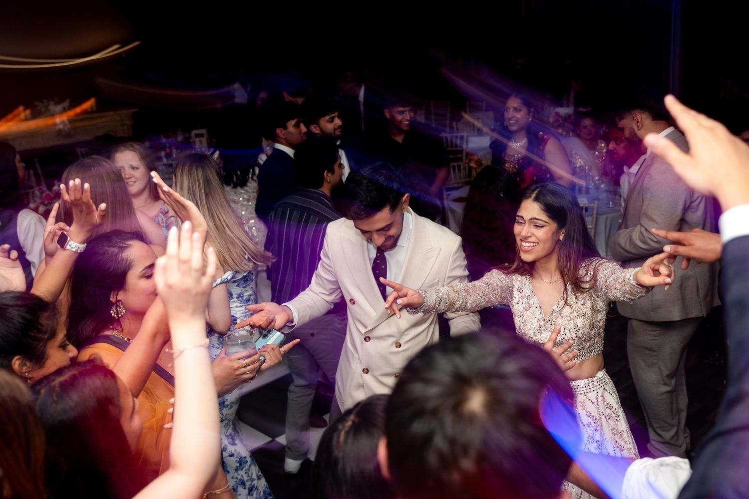 Wedding guests dance and celebrate at a lively reception with colorful purple lighting.