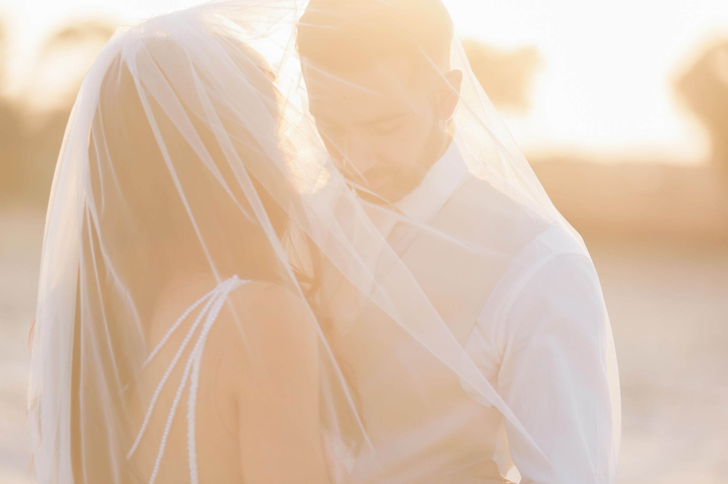 Ethereal sunlit wedding couple embraces in soft focus with bride's white veil flowing in golden hour light.