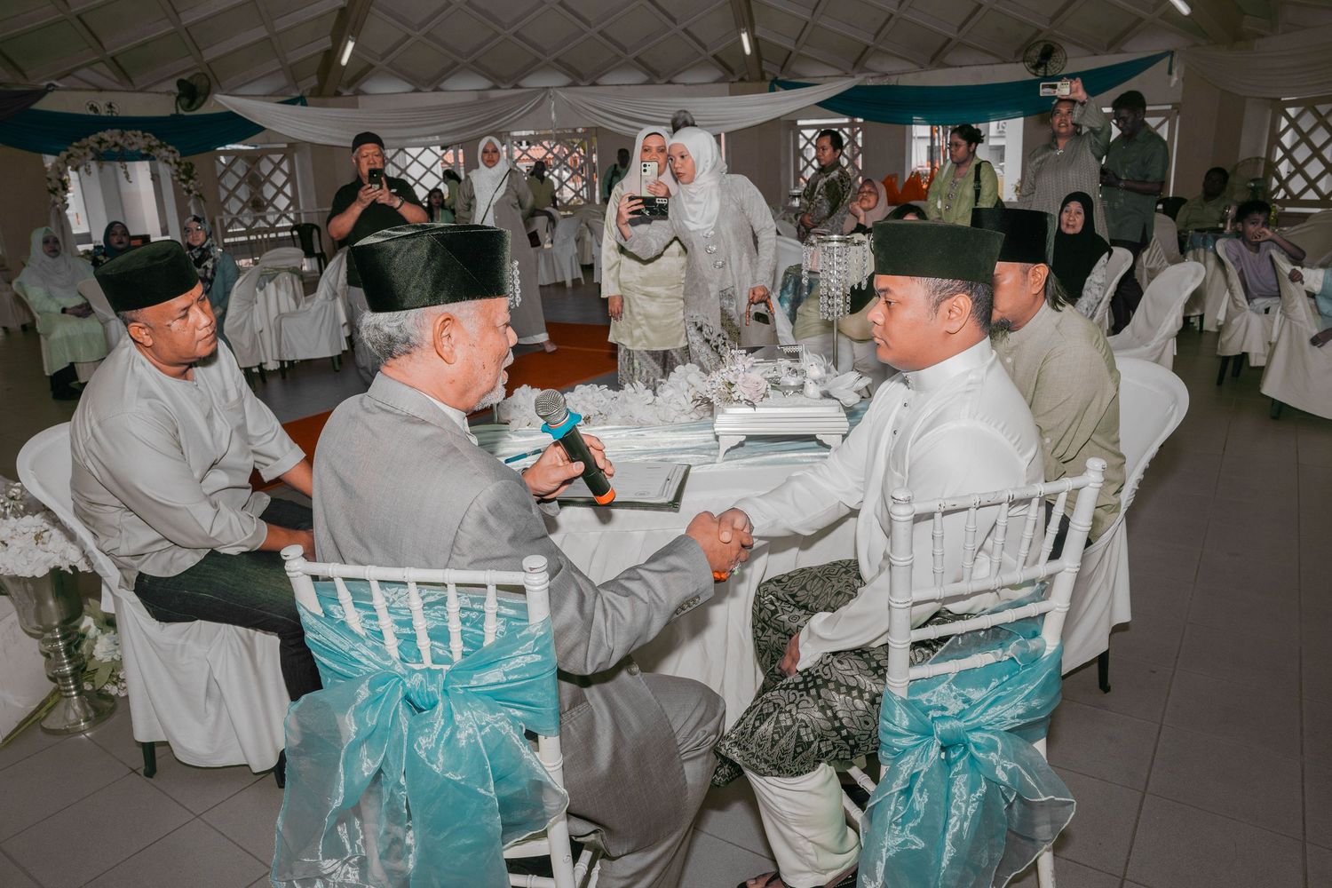 People in formal winter attire seated at round tables with turquoise chair sashes at an elegant indoor event.