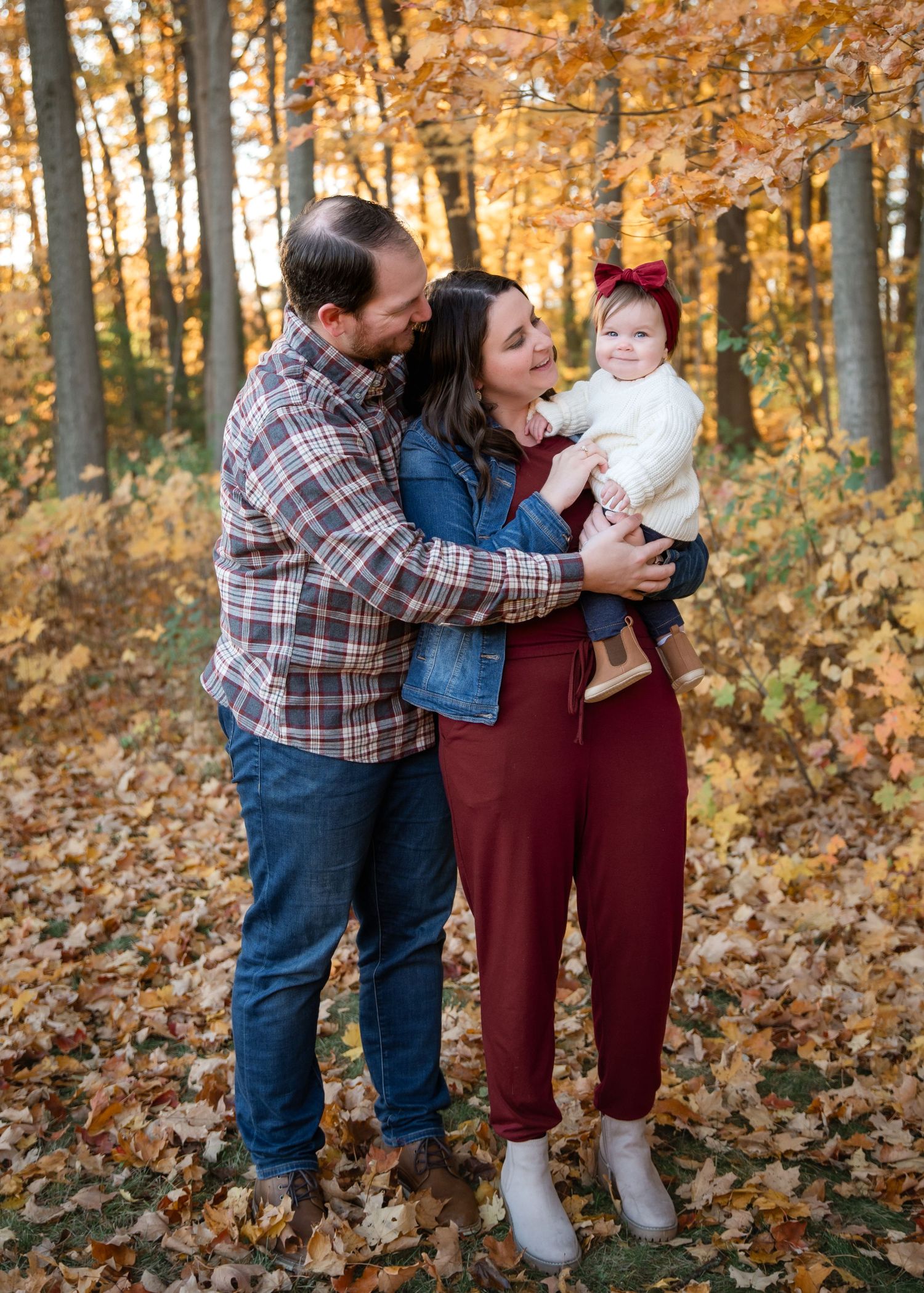 A family embraces while standing on fallen autumn leaves in a forest with golden foliage during a fall photo session.