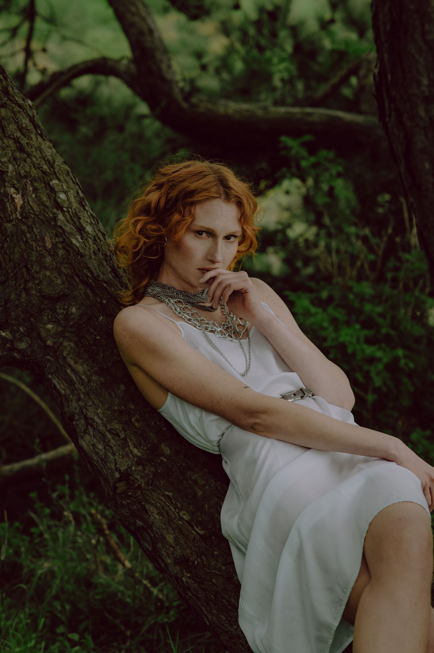 A dreamy portrait in a forest setting shows a person in white clothing resting thoughtfully against a tree trunk.