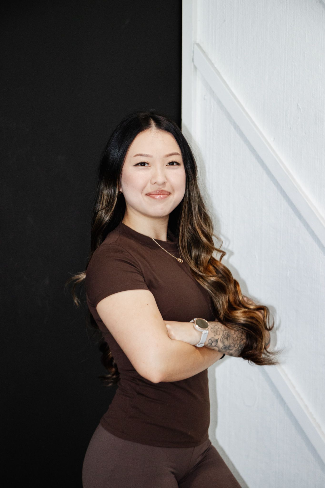 Woman with long wavy hair, wearing a brown shirt and smartwatch, stands with arms crossed, smiling confidently against a split b&w wall