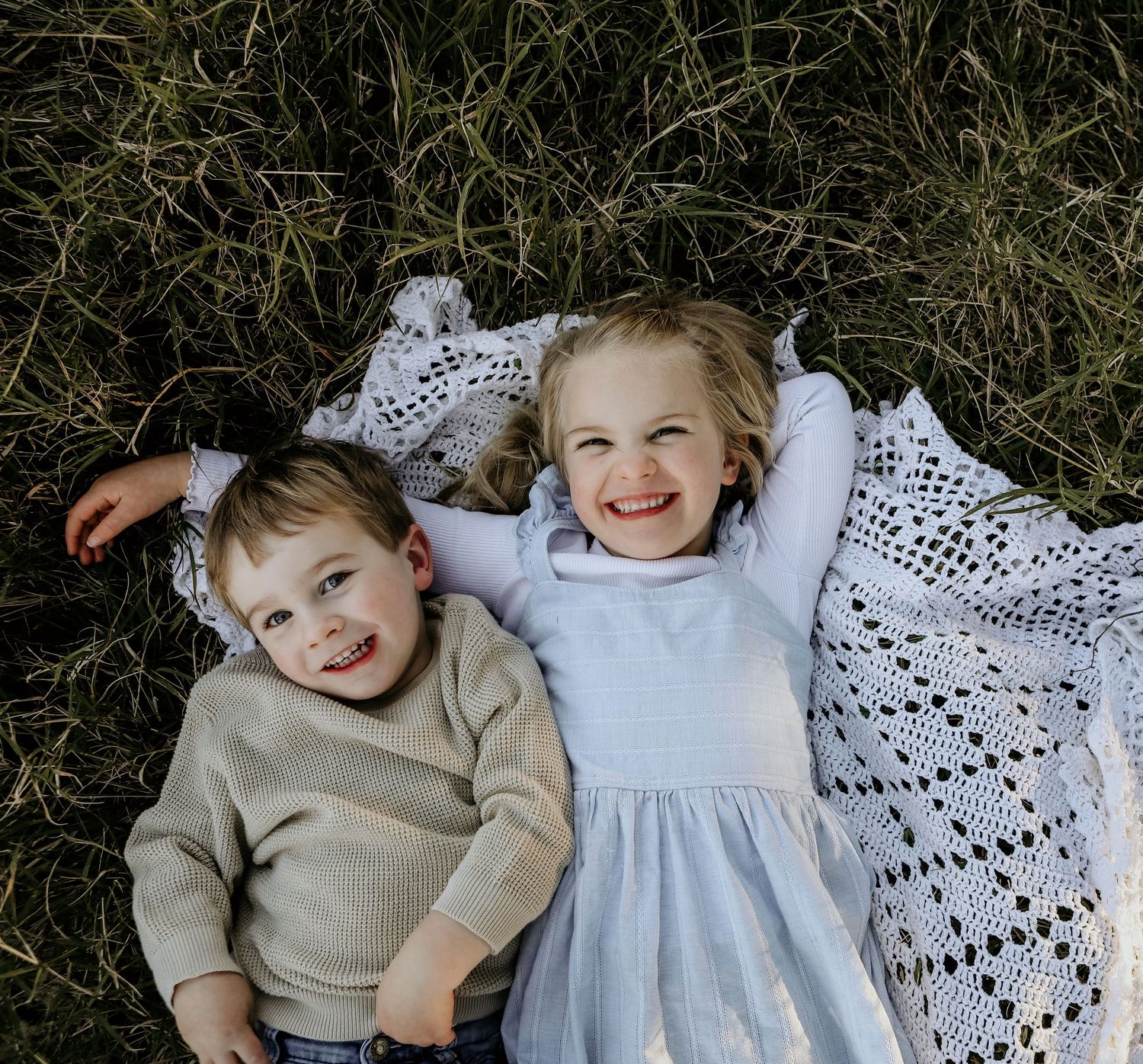 Two children lying together on a crocheted blanket in the grass during an outdoor photo session.