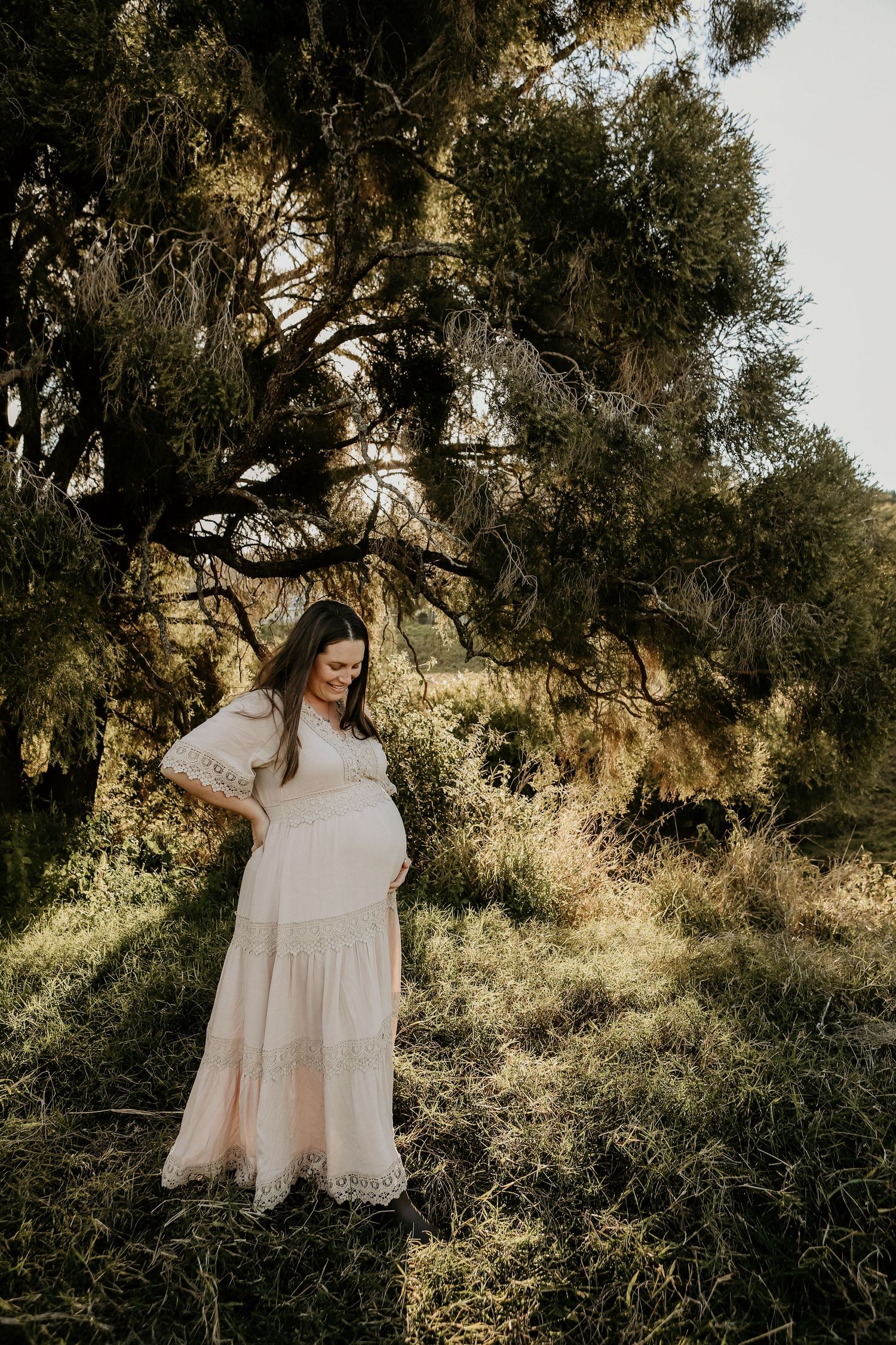 A pregnant woman stands in a flowing cream-colored dress under Spanish moss-draped trees at golden hour.