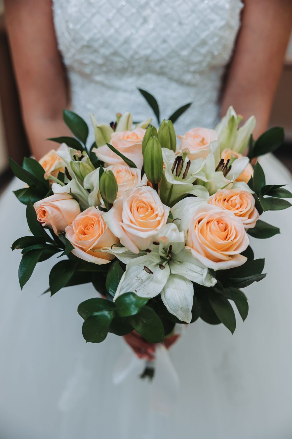 A beautiful bridal bouquet of peach roses and white lilies with dark green foliage against a white wedding dress.