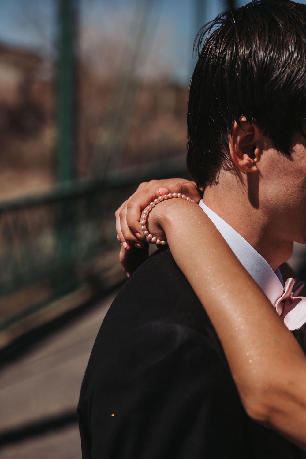 Close-up view of an embrace showing delicate pearl bracelet and formal black suit in warm sunlight.