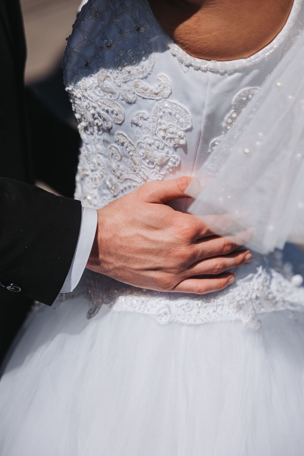 Close-up of a groom's hand on bride's waist showing intricate lace and beading details of a white wedding dress.