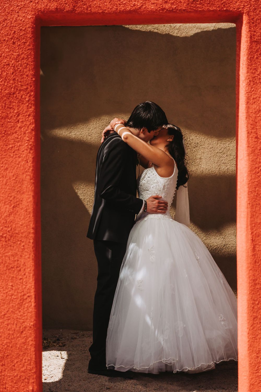 Romantic newlywed couple embraces against vibrant coral-colored wall in a series of artistic wedding portraits.