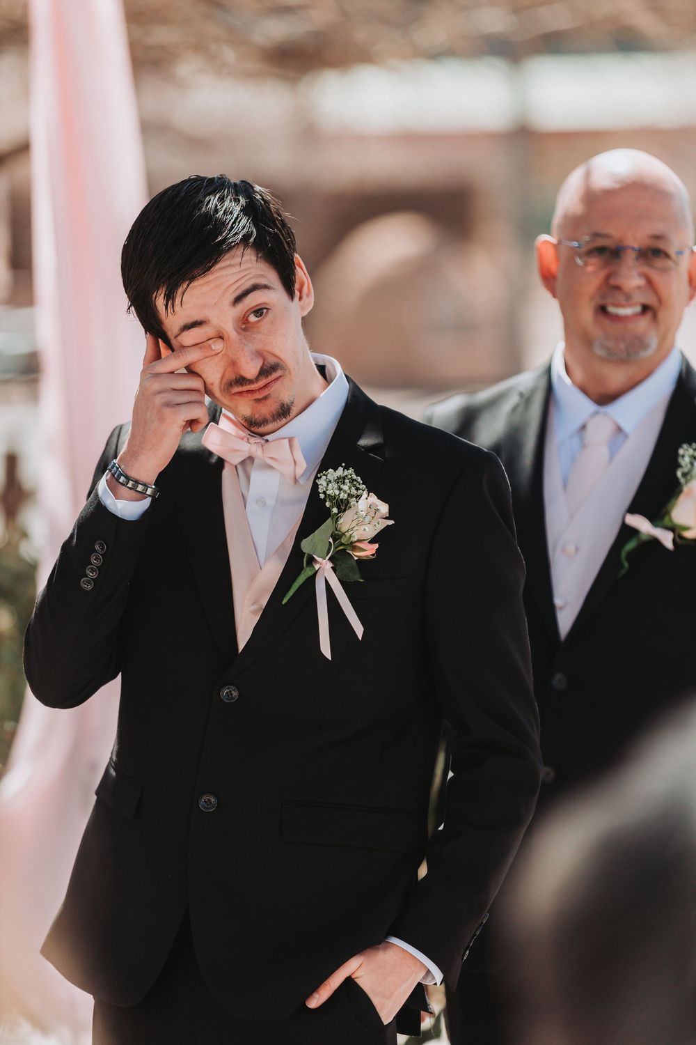 Groomsman shows emotional reaction during outdoor wedding ceremony, wearing black suit with boutonniere.