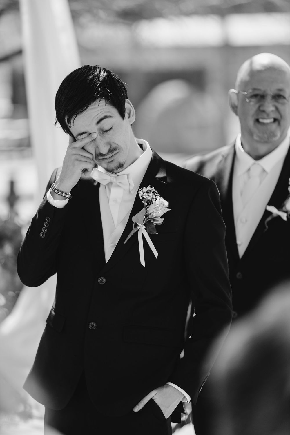 Black and white wedding photo of emotional groom wiping tears during ceremony as groomsman smiles in background.