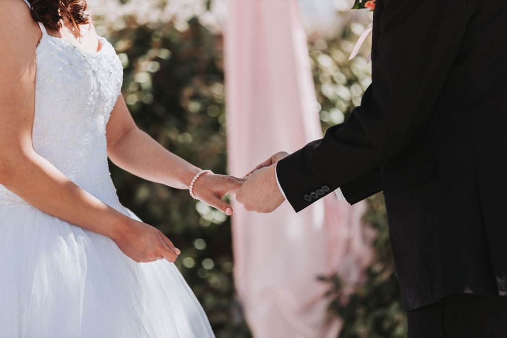 A bride in a white wedding dress holds hands with someone wearing a black suit during an outdoor ceremony.